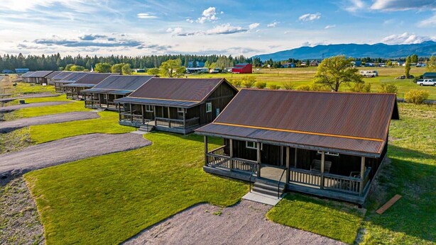 A row of wooden cabins with metal roofs, set in a grassy area with a mountain backdrop. - The Kabins - Camp Misery (Columbia Falls)