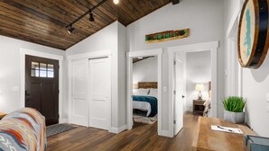 A spacious room with a wooden ceiling, white walls, and a bed with a colorful blanket. There is a door leading to another room, a small table with a lamp, and a plant on the right side. - The Kabins - Camp Misery (Columbia Falls)