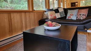 A cozy room with a wooden interior, a black leather sofa with decorative pillows, a small table with a bowl of candles, and large windows offering a view of the forest.