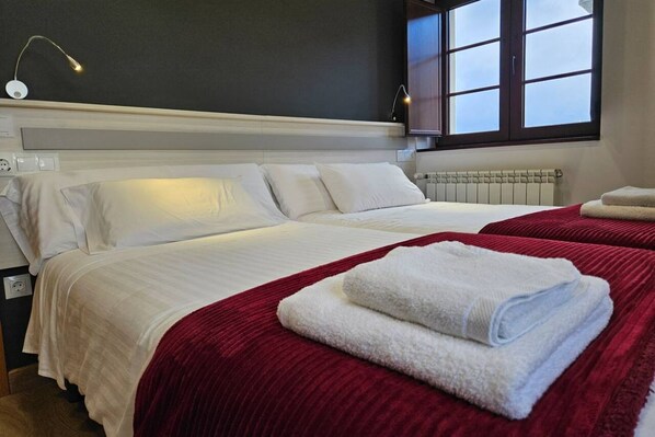 A hotel room with two beds, a red blanket, white towels, a window with a view, and a wall-mounted lamp.