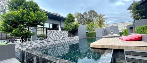 A modern outdoor swimming pool with a tiled deck, surrounded by greenery and a contemporary building.