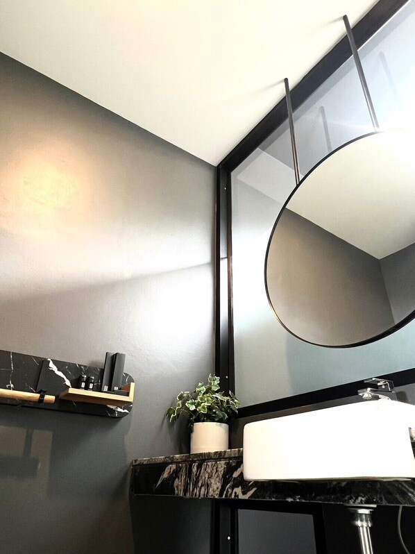 Un baño moderno con encimera de mármol negro, lavabo blanco, un espejo grande y una pequeña planta en maceta.