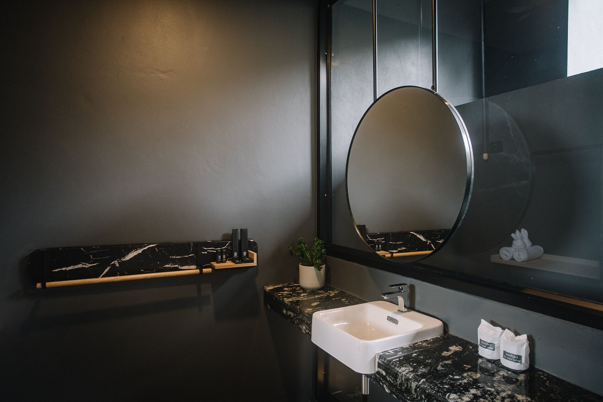 A modern bathroom with a black marble countertop, a white sink, and a large mirror. There is a shelf with toiletries and a potted plant.