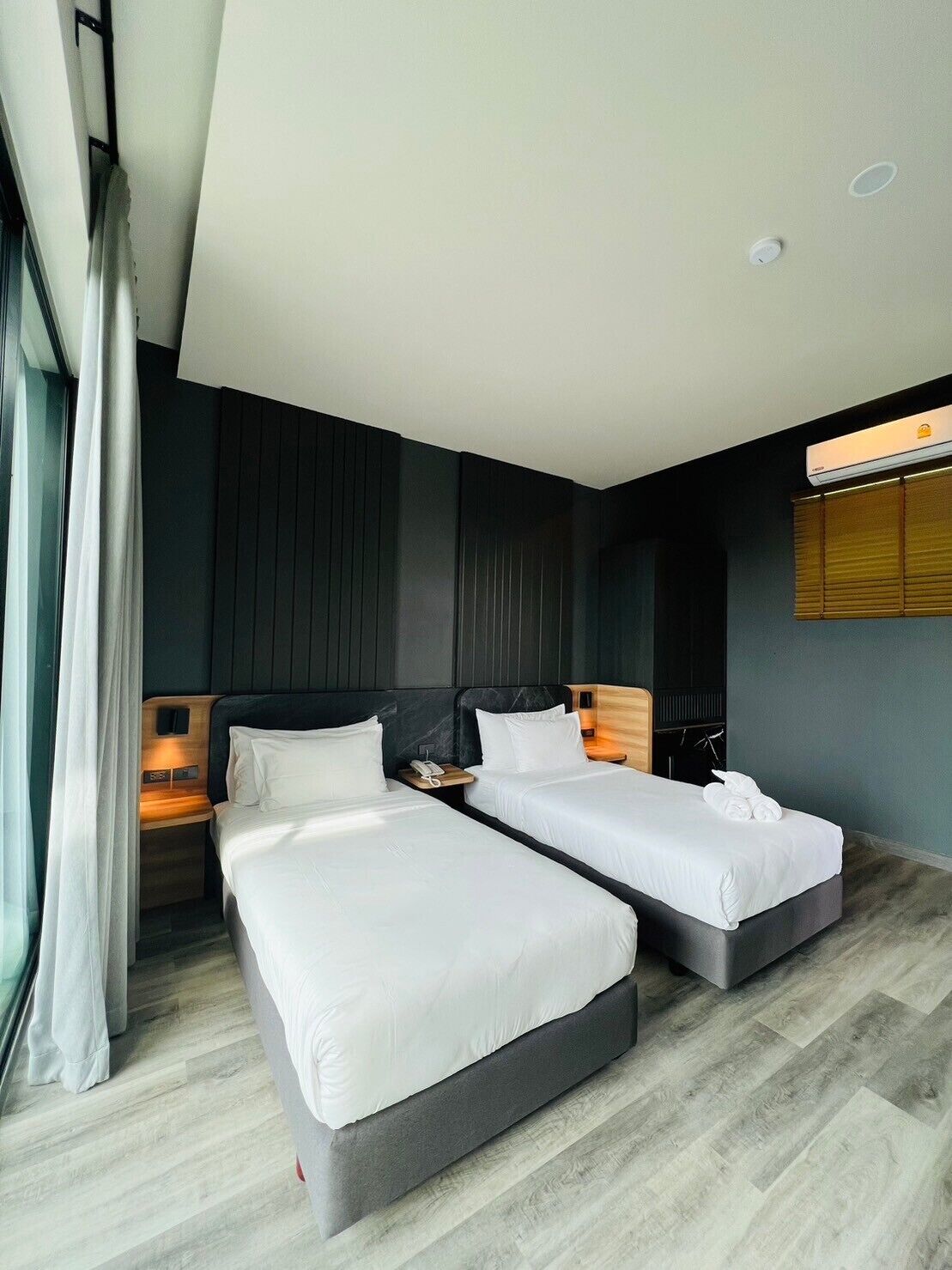 Habitación de hotel moderna con dos camas individuales, una mesita de noche de madera con un teléfono, aire acondicionado en la pared y una ventana con cortinas.