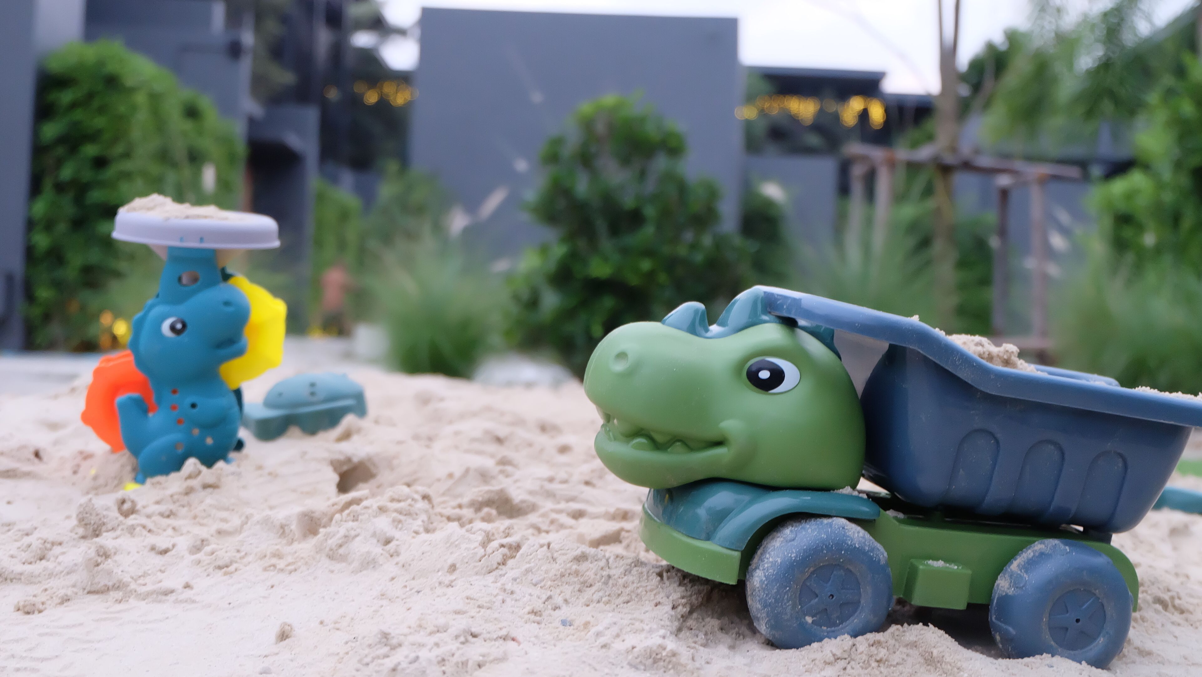 Un camión volquete de juguete sobre arena, con un dinosaurio de juguete y un coche de juguete al fondo.