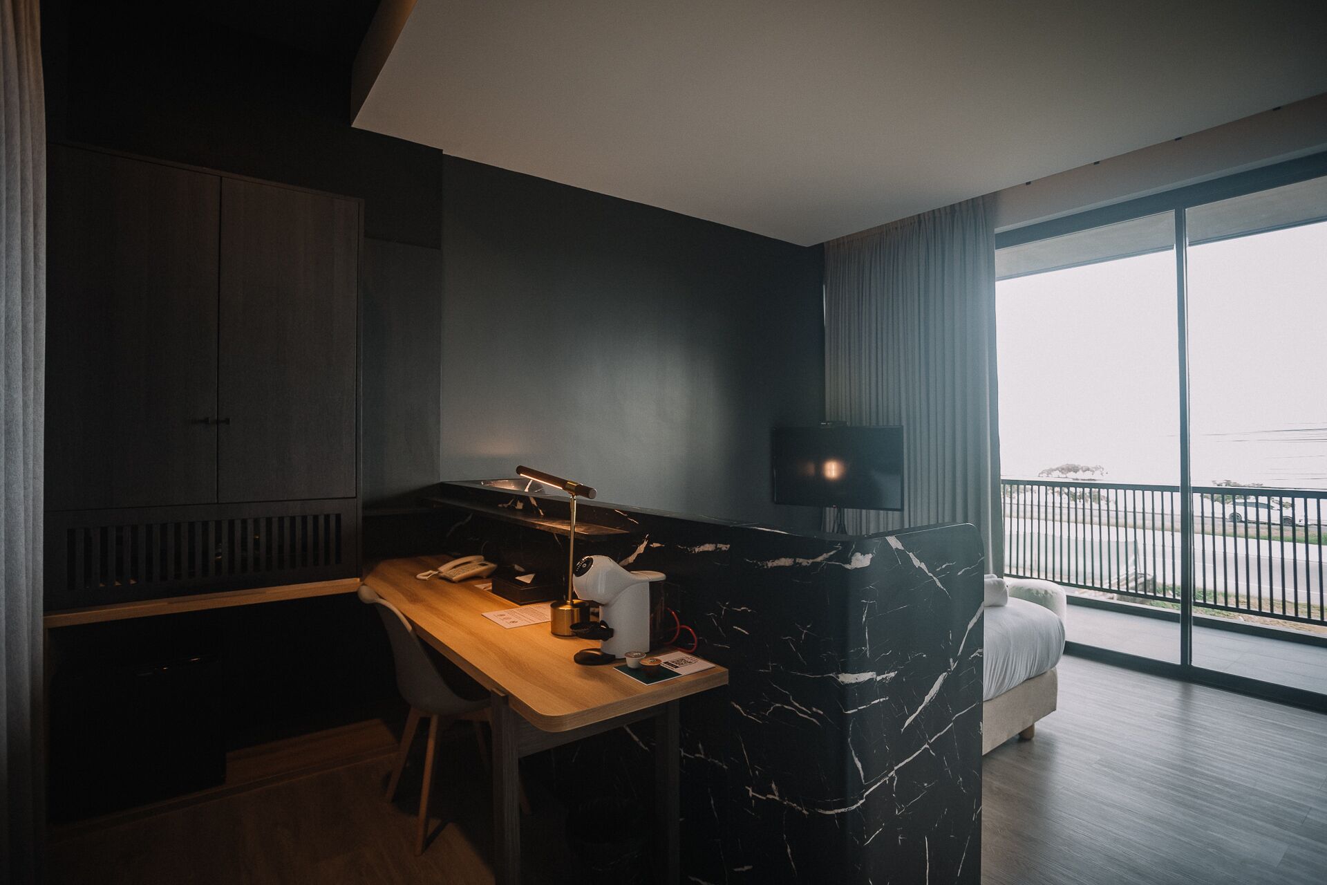 Quarto de hotel moderno com escrivaninha de madeira, parede de destaque em mármore preto, televisão de tela plana e varanda com vista.