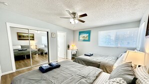 A bedroom with two beds, a ceiling fan, a window with blinds, and a door leading to another room.
