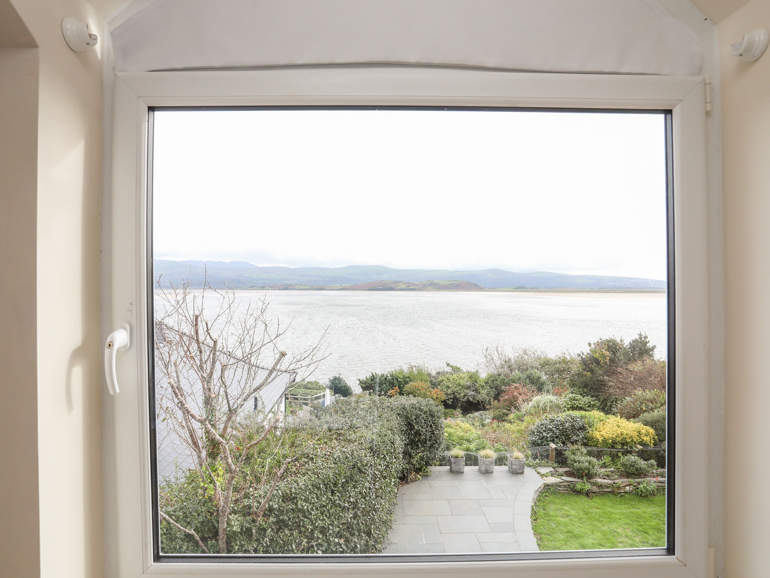 Vista desde una ventana de una habitación con puerta, un jardín con sendero y una masa de agua a lo lejos.