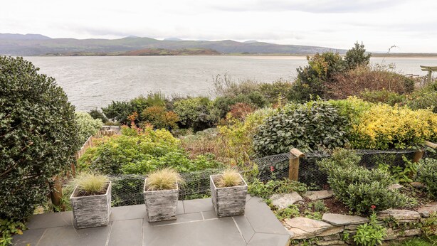 Un patio con piso de piedra, rodeado de diversas plantas y arbustos, con vista a una masa de agua y colinas distantes.