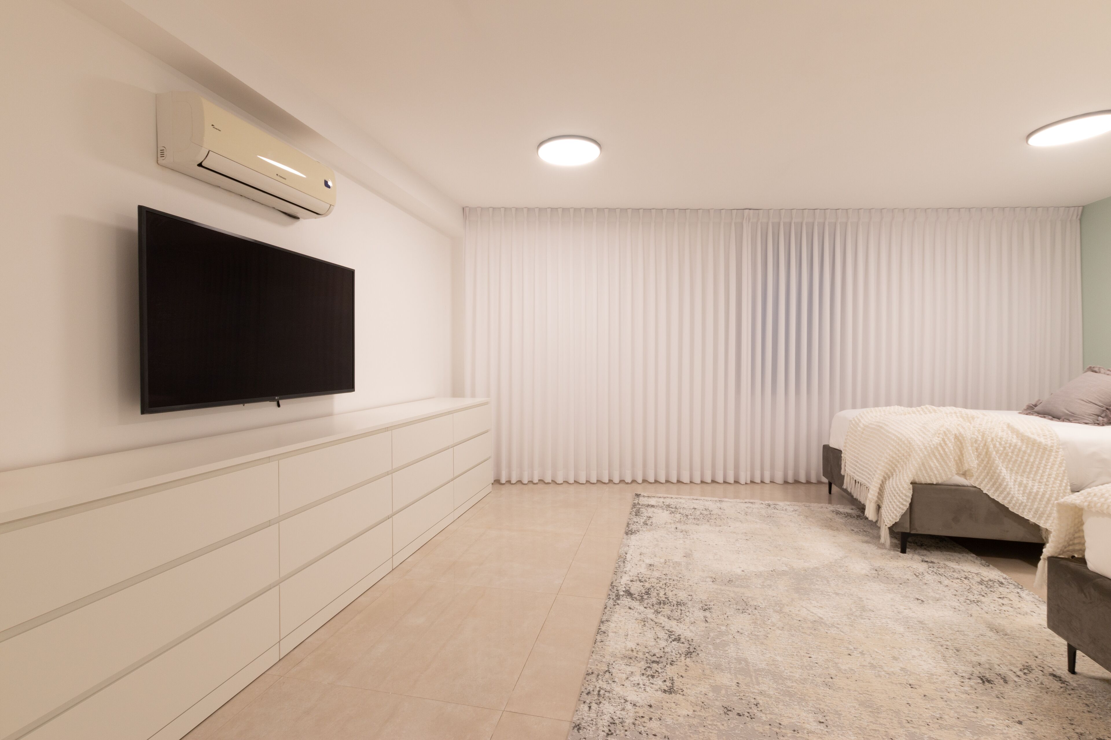 A modern bedroom with a flat-screen TV mounted on the wall, a bed with a white cover, a large window with vertical blinds, and a beige carpet.