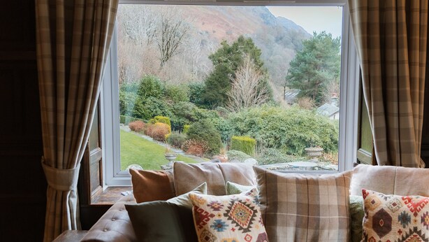 A view from a window with curtains, a sofa with patterned cushions, and a garden with trees and a lawn. - Grey Walls (Penrith)