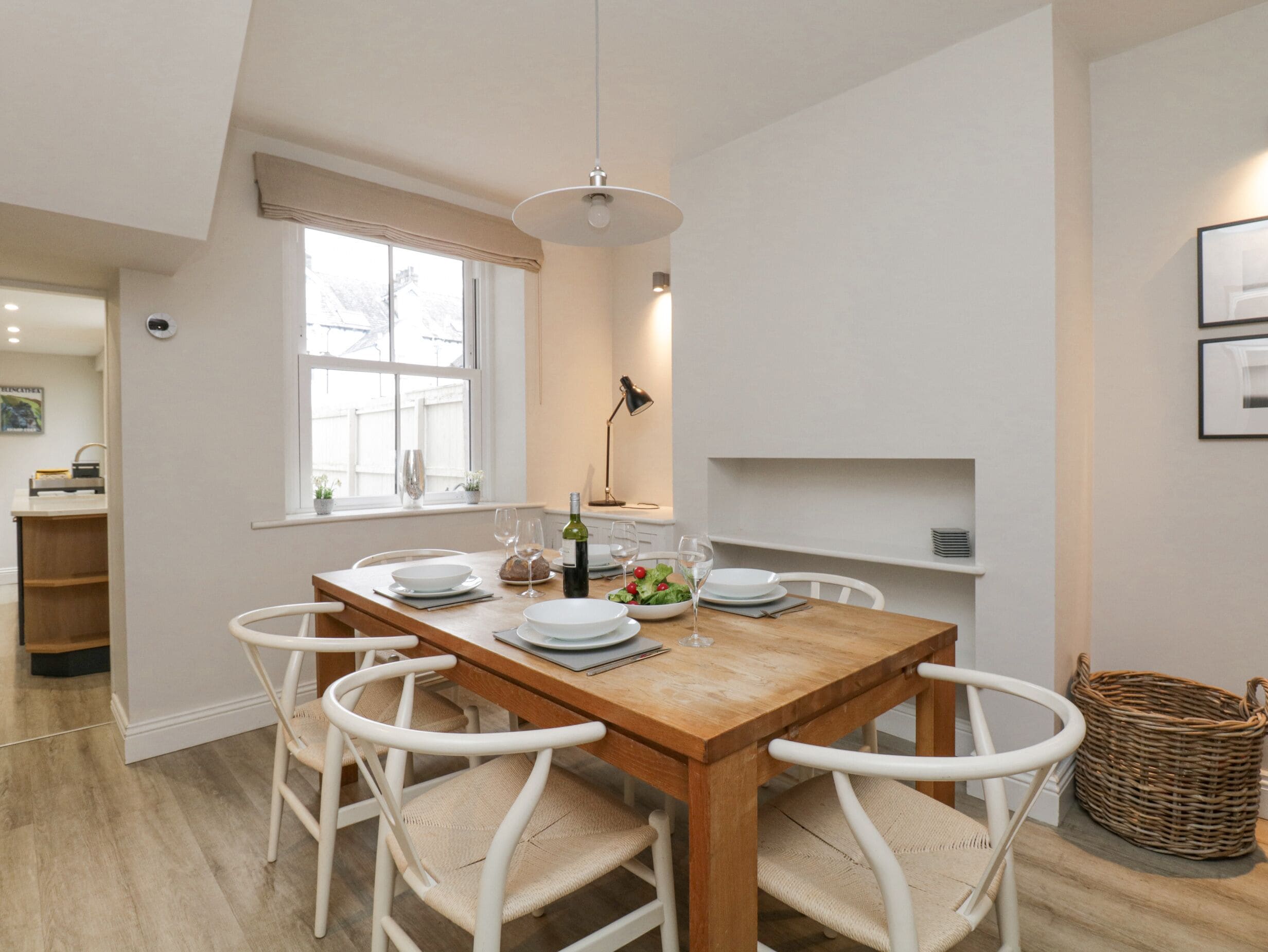Un coin repas avec une table en bois et des chaises blanches, une fenêtre avec des rideaux et une lampe fixée au mur.