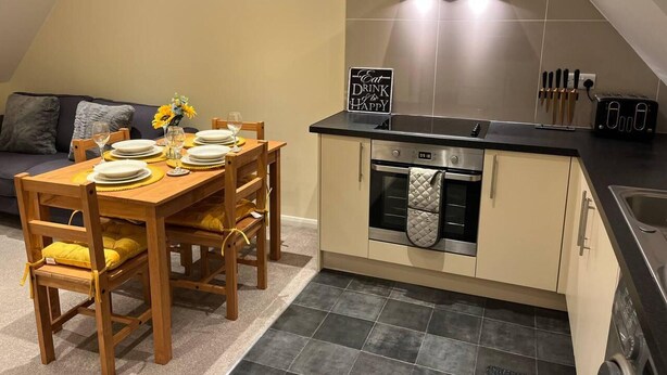 A dining area with a wooden table set for four, a kitchen with a stove and oven, and a sofa in the background. - Central Location 2-bed Apartment in Maidstone (Maidstone)