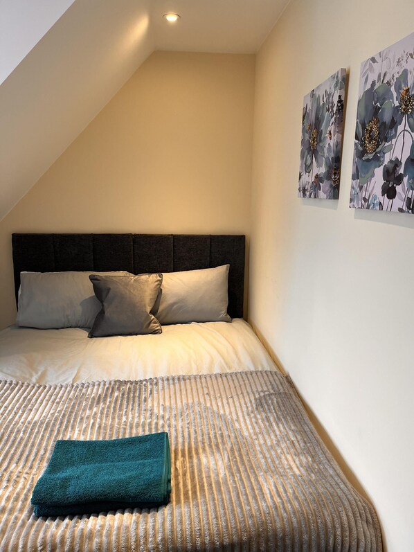 Un dormitorio con techo inclinado, una cama con cabecera oscura, dos almohadas, un plaid verde aguamarina y dos cuadros florales.