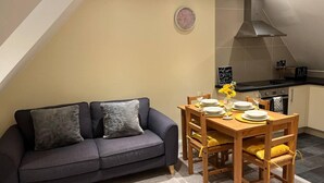 A cozy living room with a dark grey sofa, a wooden dining table set with plates and glasses, and a kitchen with a modern stove and cabinets. - Central Location 2-bed Apartment in Maidstone (Maidstone)