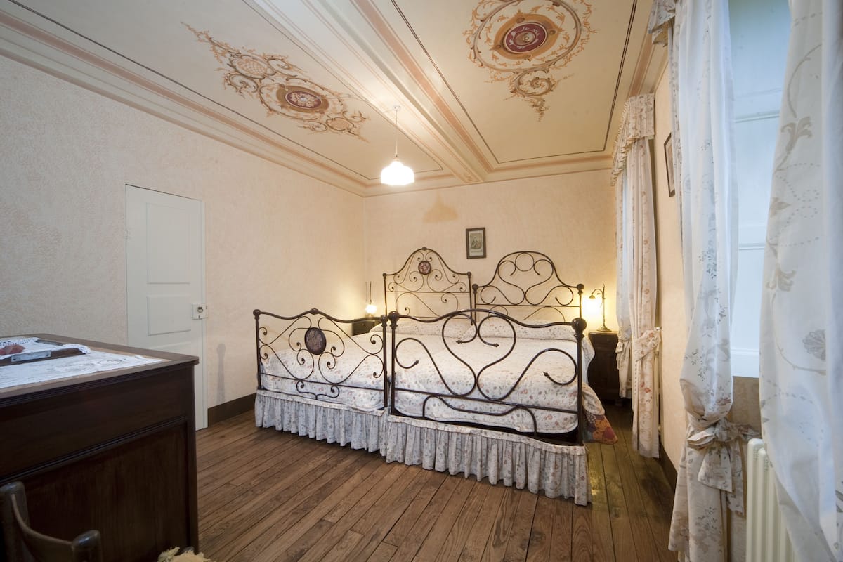 A room with a wooden floor, a bed with ornate metal headboard, a desk, and a door.