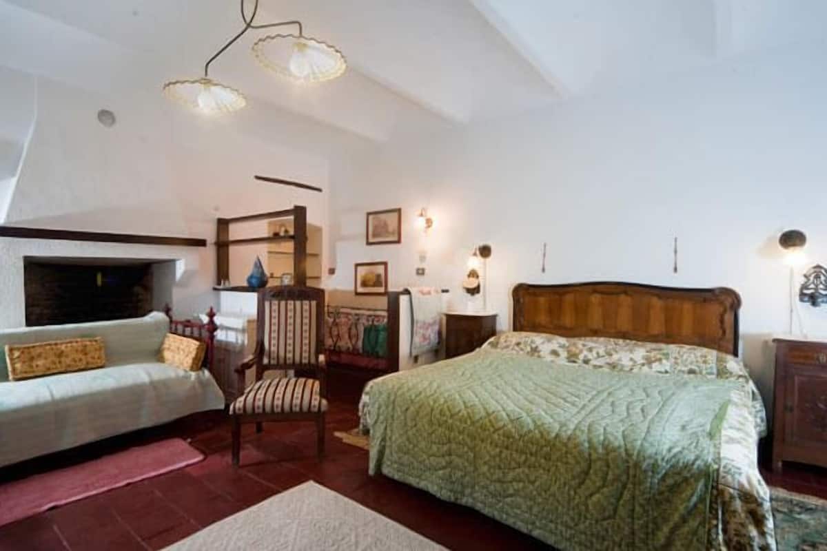 A bedroom with a bed, a chair, a sofa, a fireplace, and wall-mounted lights.