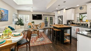 Un salon spacieux comprenant un canapé, des fauteuils, une table à manger et un téléviseur. Il y a une cuisine avec un coin repas, un ventilateur de plafond et une horloge.