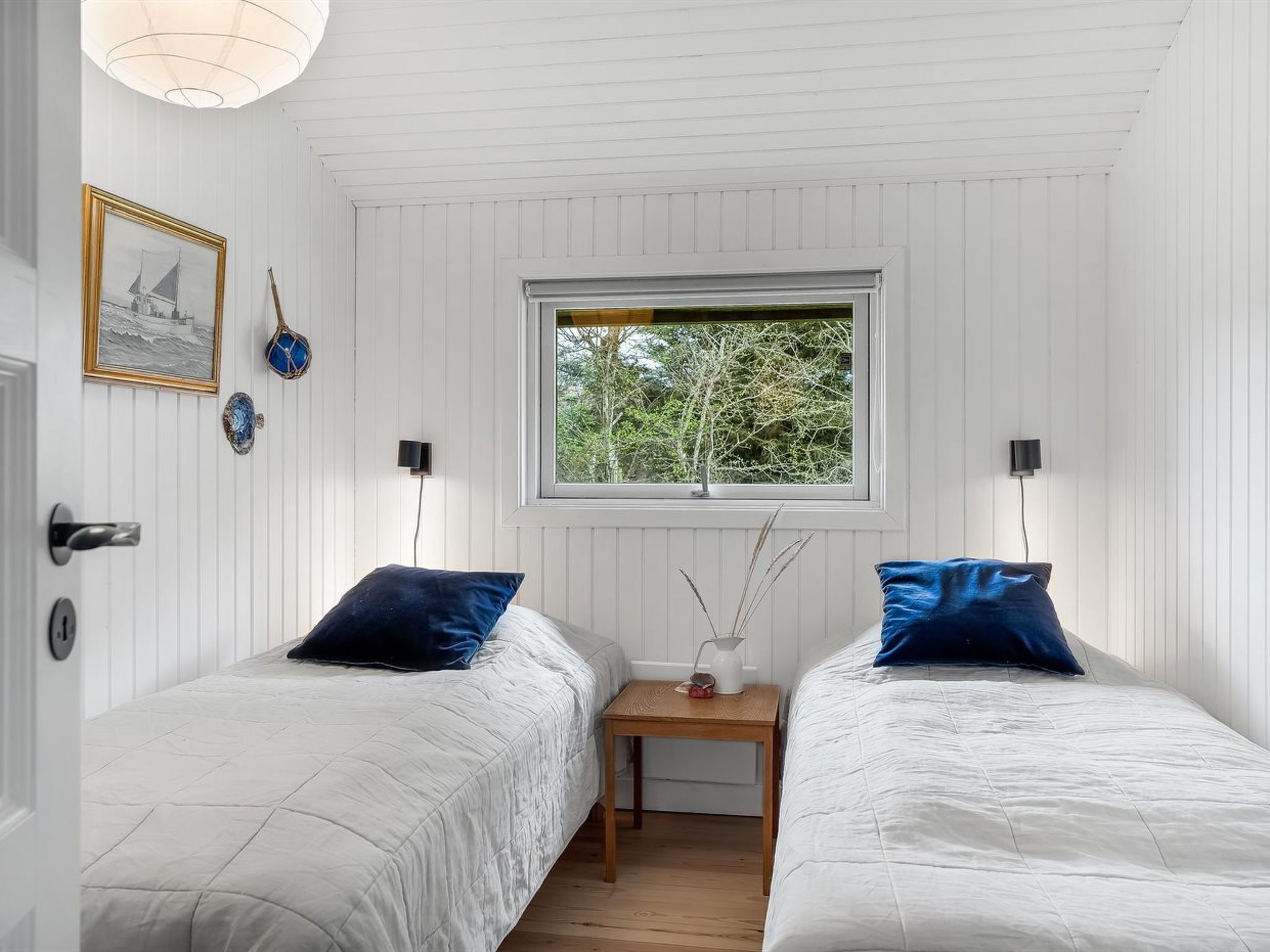 Une chambre avec deux lits, une fenêtre donnant sur des arbres, une petite table avec un vase et des lampes fixées au mur.