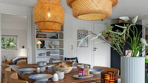 Un coin repas avec une table en bois dressée pour quatre personnes, entourée de chaises en osier. Deux grandes lampes suspendues en osier sont accrochées au-dessus de la table.