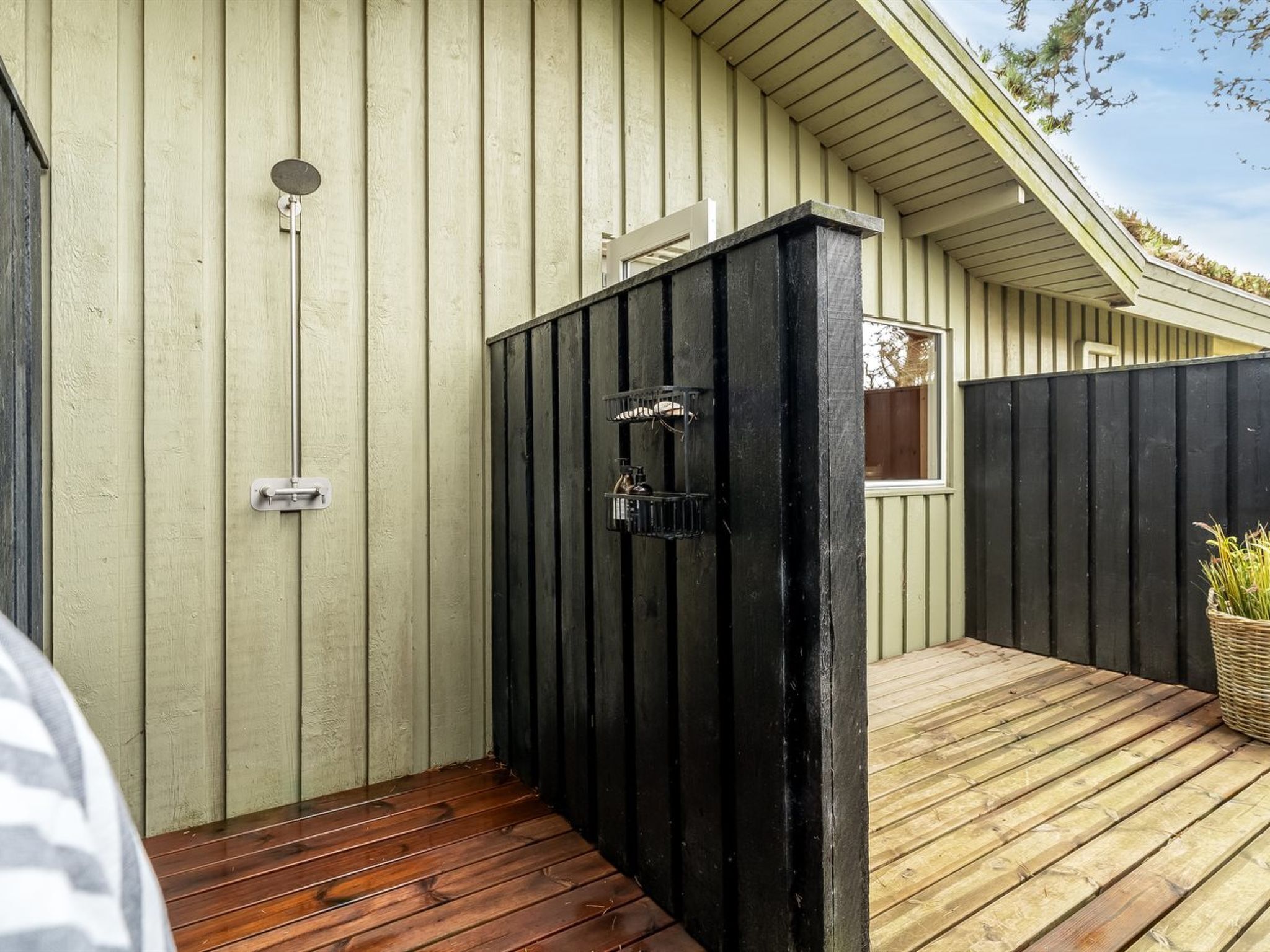 Une terrasse en bois agrémentée d’un paravent noir pour plus d’intimité, d’un robinet en métal et d’une fenêtre donnant sur le ciel.