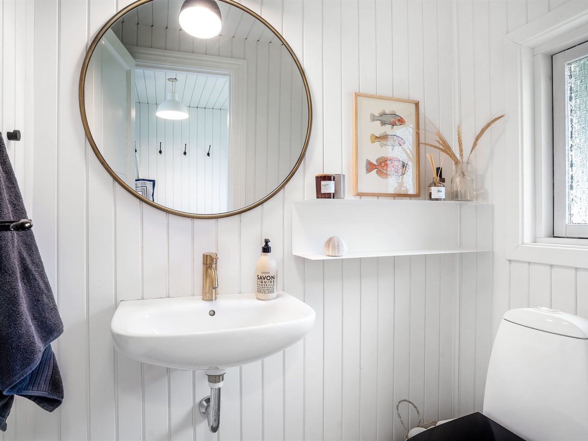 Une salle de bain avec un lavabo blanc, un miroir rond et des toilettes. On y trouve un porte-serviettes sur lequel est accrochée une serviette.