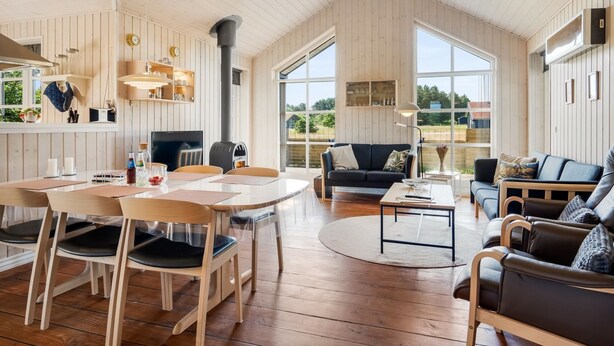 A spacious living area with a high ceiling, wooden flooring, a dining table with chairs, a sofa, and a coffee table. There are large windows providing natural light and a view of the outdoors.