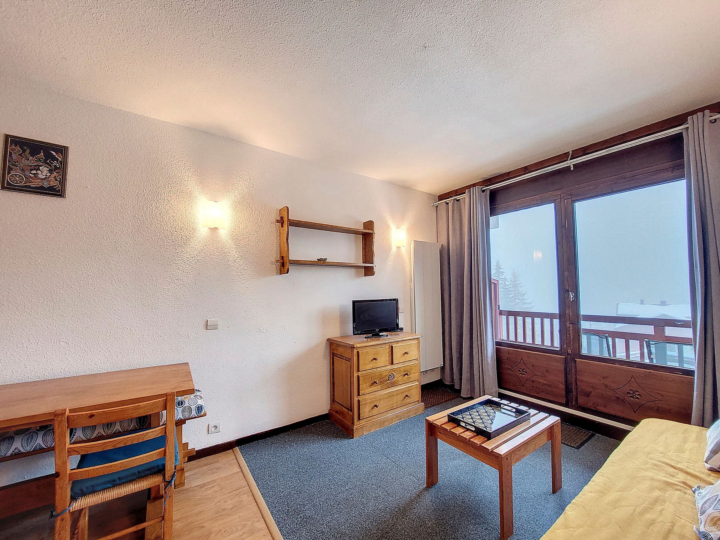 A furnished room with a wooden desk, a television, a small table with a board game, and a balcony with a view.