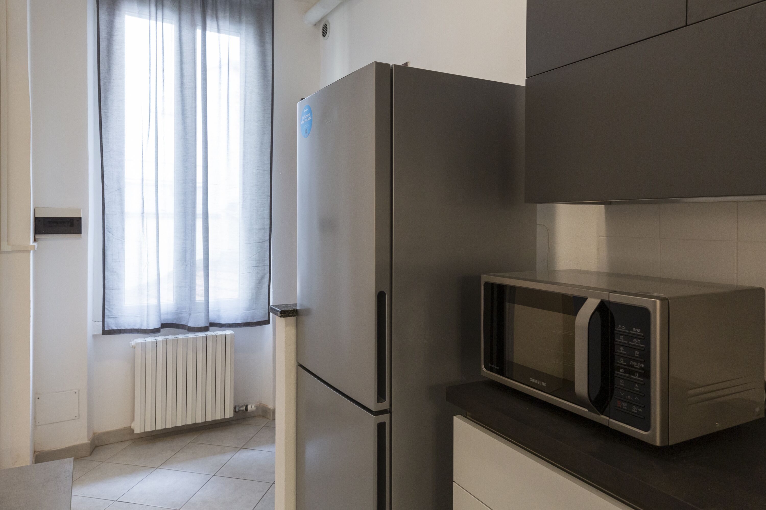 Une cuisine moderne équipée d’un réfrigérateur en acier inoxydable, d’un four à micro-ondes et d’une fenêtre agrémentée de rideaux légers.