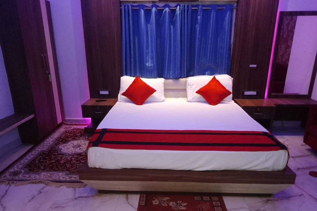 A bedroom with a large bed, two nightstands, a red and black bed runner, a patterned rug, and a blue curtain.