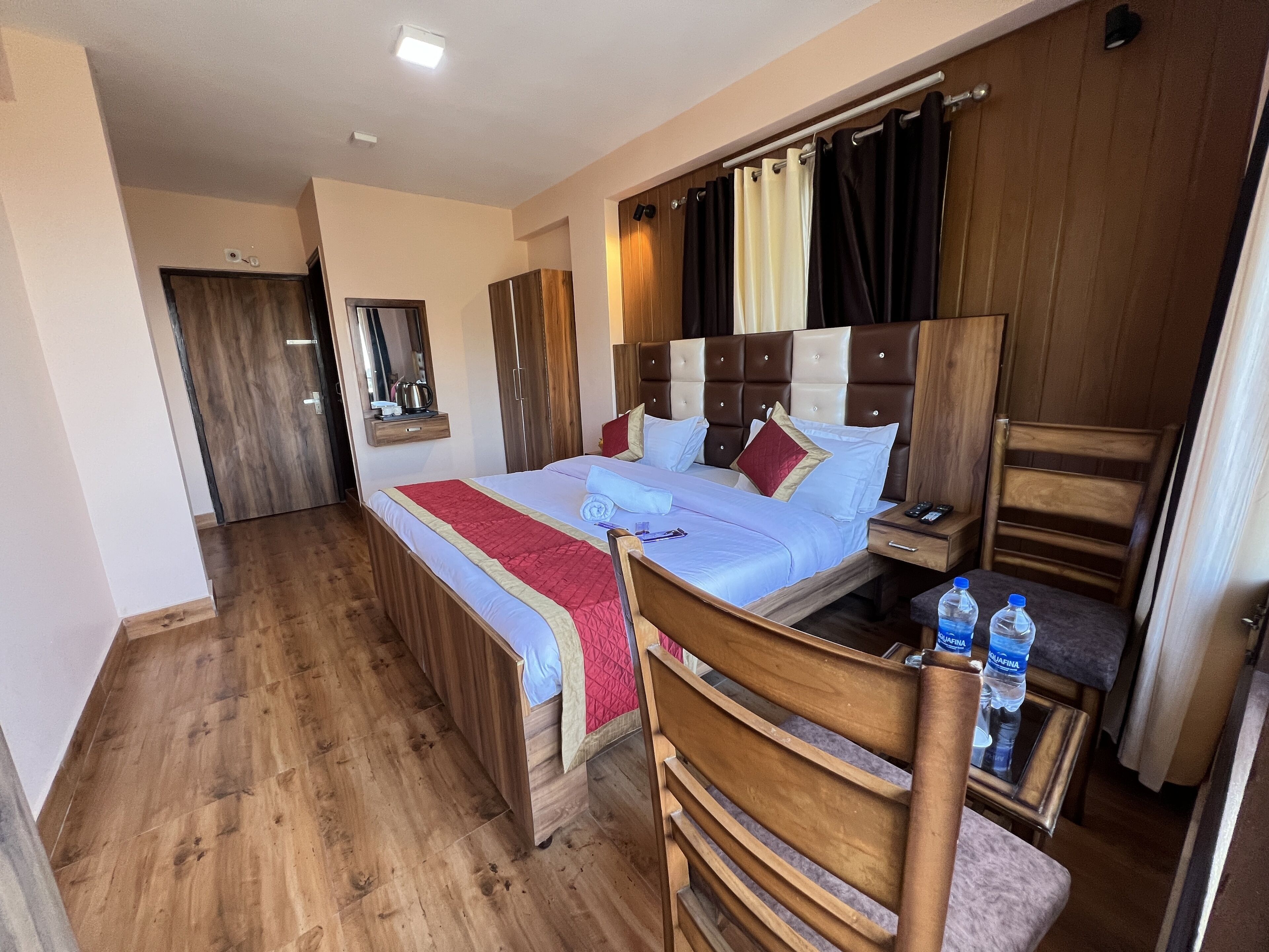 A hotel room with a wooden floor, a large bed with a headboard, a nightstand, a chair, and a small table with water bottles.