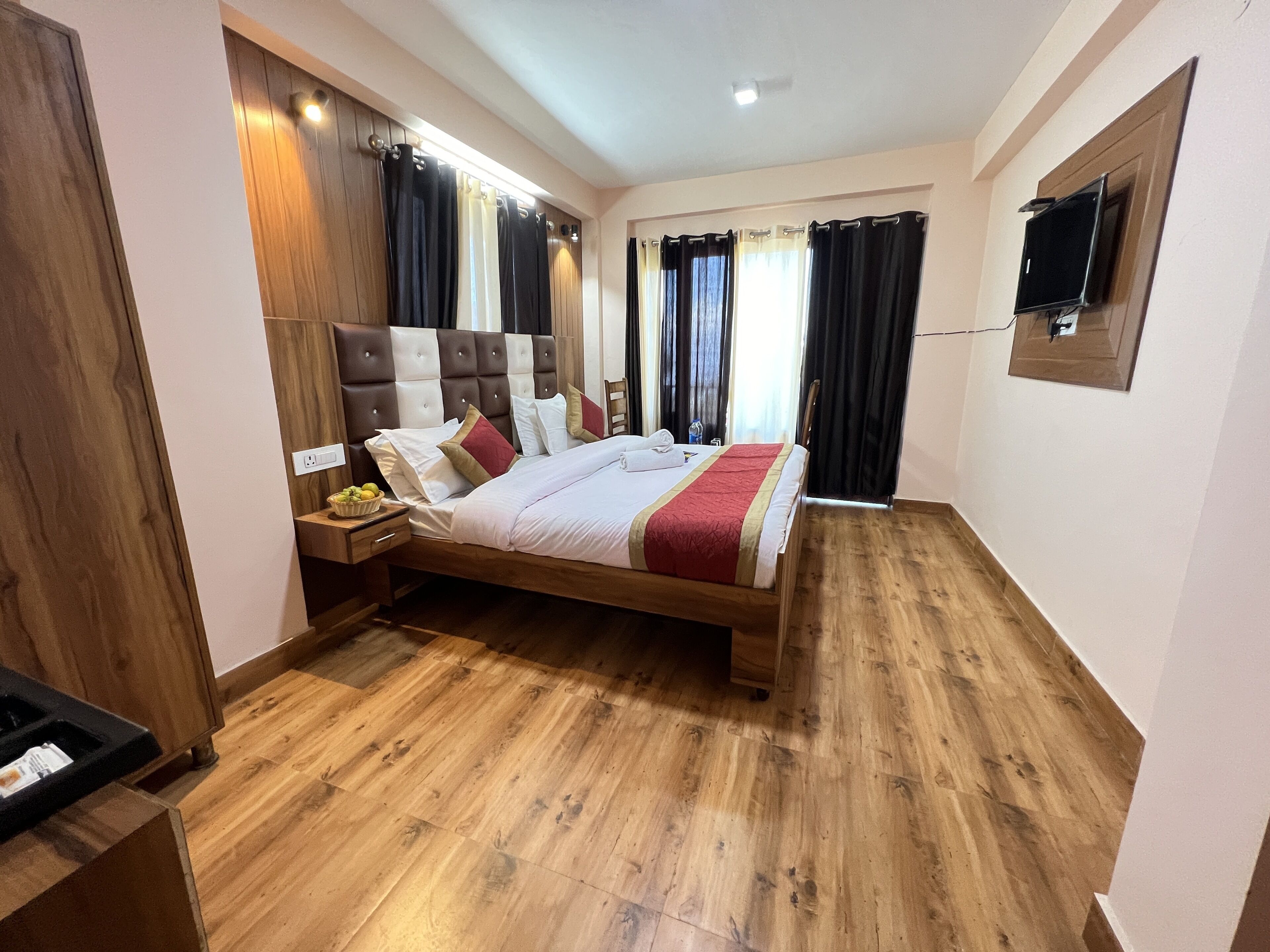 A modern hotel room with a wooden floor, a large bed with multiple pillows, a nightstand with a fruit bowl, and a TV mounted on the wall.