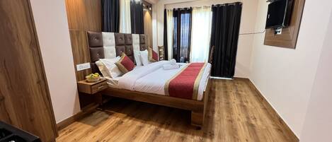 A modern hotel room with a wooden floor, a large bed with multiple pillows, a nightstand with a fruit bowl, and a TV mounted on the wall.
