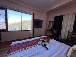 Habitación de hotel con una cama grande, un televisor montado en la pared, una ventana con cortinas, una mesita con un frutero y una silla.