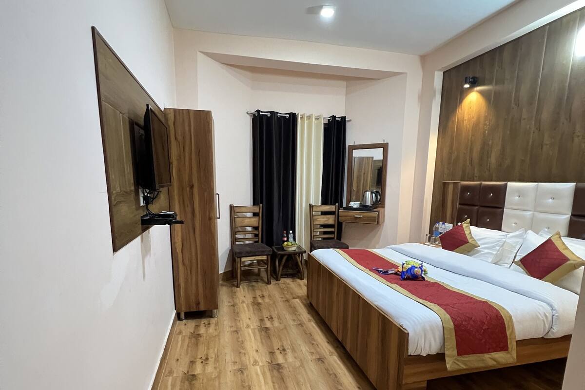 Habitación de hotel con suelo de madera, cama con colcha roja y blanca, armario de madera, una mesita con una silla, un espejo y televisor montado en la pared.