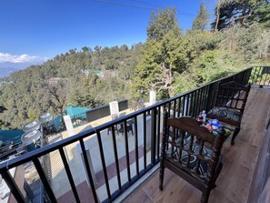 A balcony with a wooden chair and a table, overlooking a mountainous area with trees and houses. - Canadian Cottage Dalhousie  (Dalhousie)
