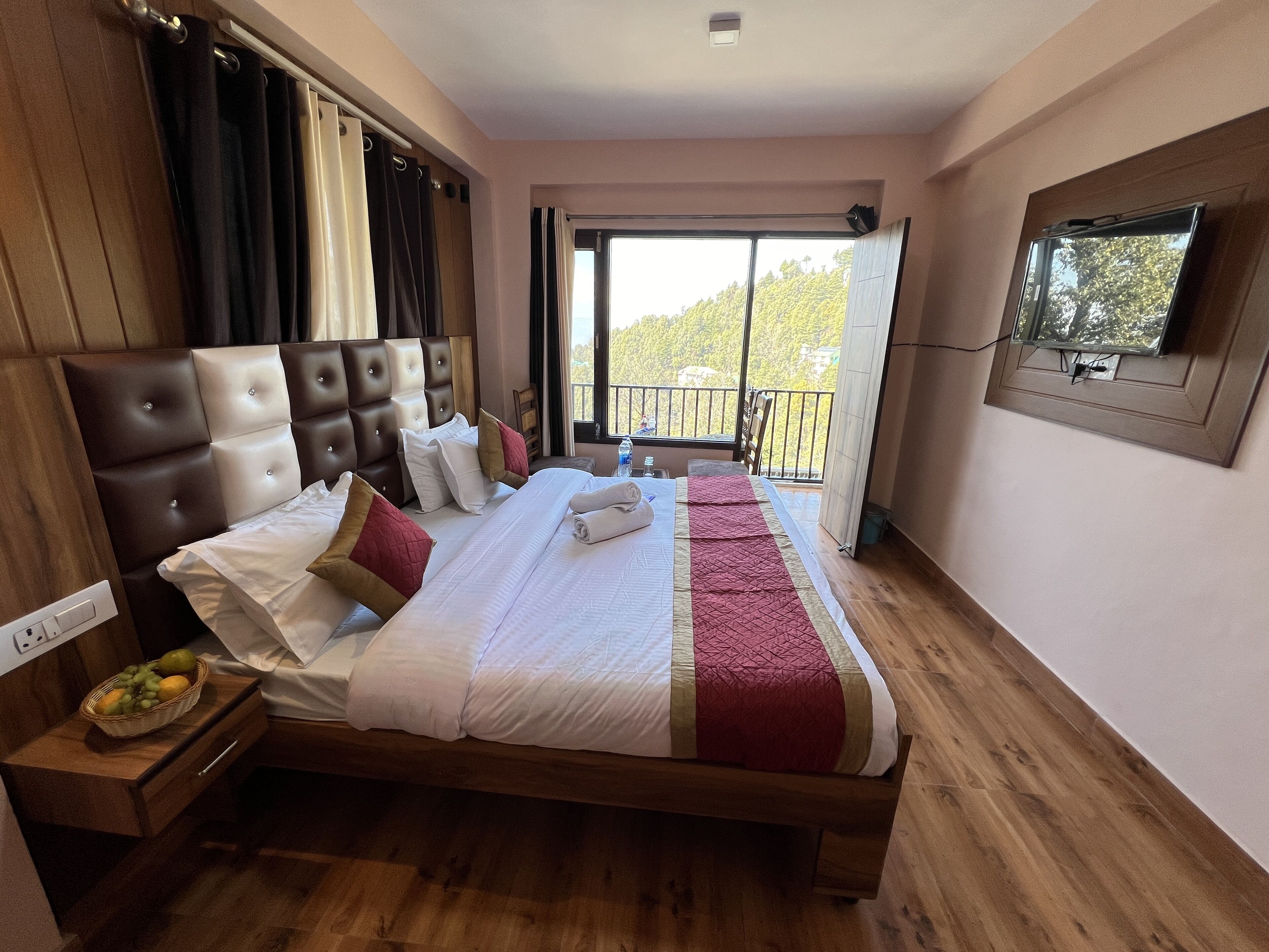 A hotel room with a large bed, a wooden headboard, a TV mounted on the wall, and a balcony with a view of trees.