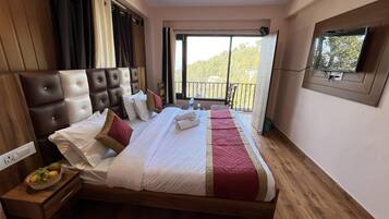 Una habitación de hotel con una cama grande, cabecera de madera, televisor montado en la pared y un balcón con vistas a árboles.