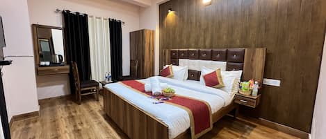 A hotel room with a wooden floor, a large bed with a headboard, a bedside table with a lamp, a mirror, and a window with curtains.