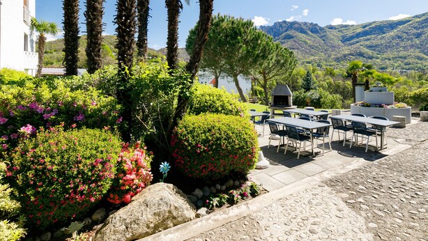 Eine Terrasse im Freien mit Tischen und Stühlen, umgeben von üppiger Vegetation und Palmen, mit Blick auf eine gebirgige Landschaft.