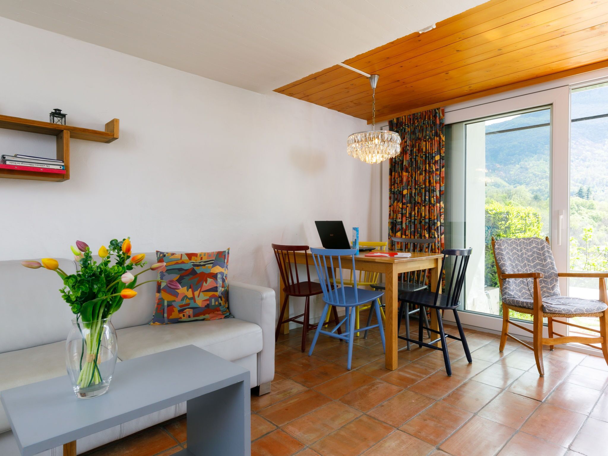 A cozy living room with a sofa, a coffee table, and a dining table with chairs. There is a laptop on the table, a vase with flowers on the coffee table, and a bookshelf with books. The room has a wooden ceiling and a large window with a view of greenery.
