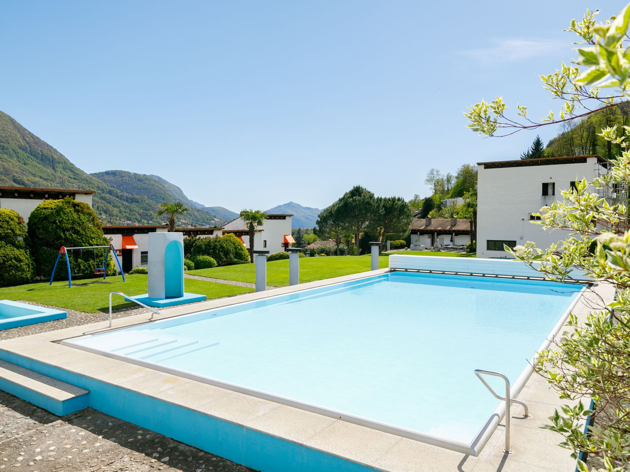 A swimming pool with a clear blue water, surrounded by a white deck and blue steps. In the background, there are buildings and a mountain.