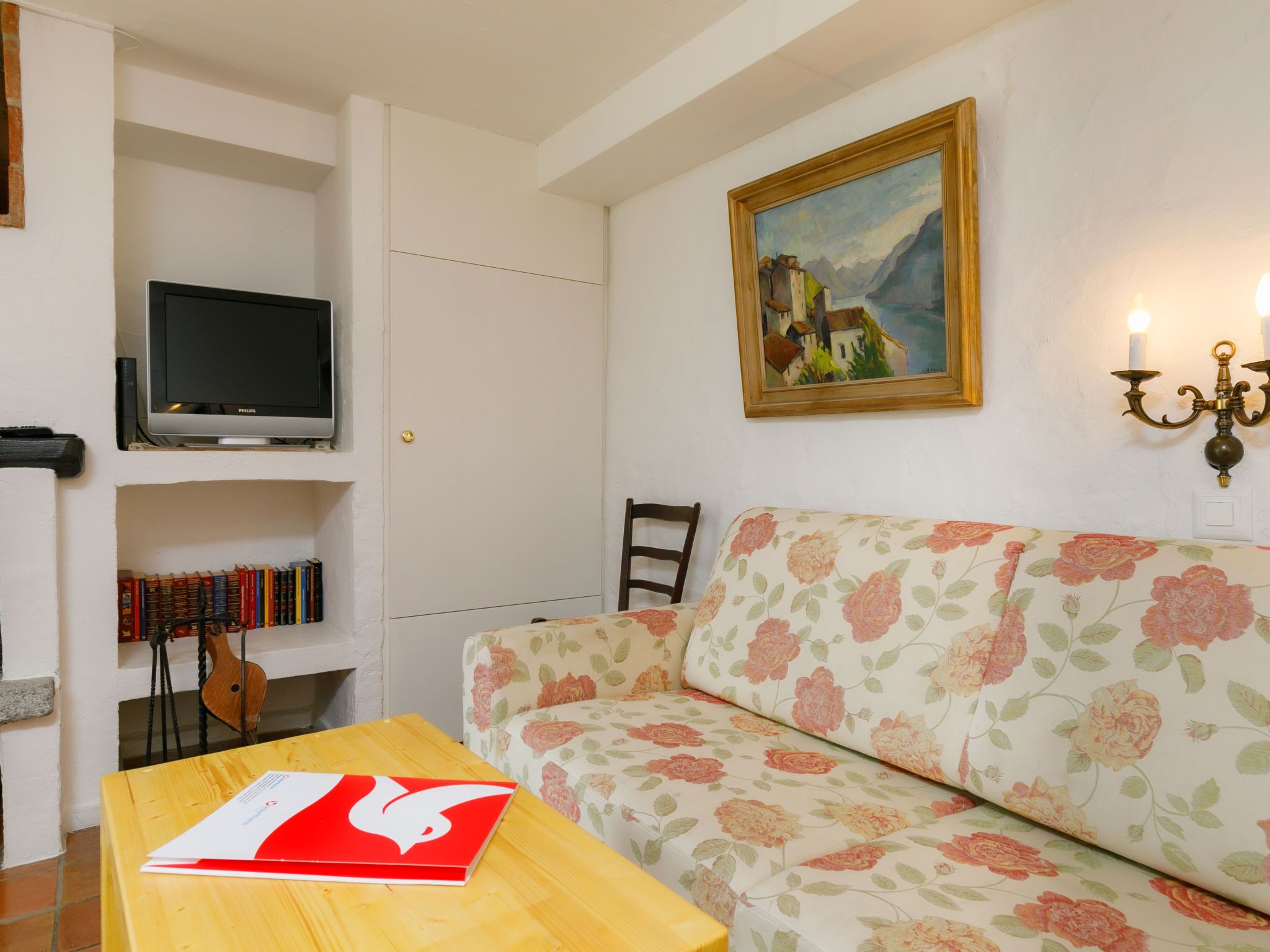 Un salon avec un canapé à motifs floraux, un téléviseur posé sur une étagère, un tableau accroché au mur et une table sur laquelle se trouve un livre.