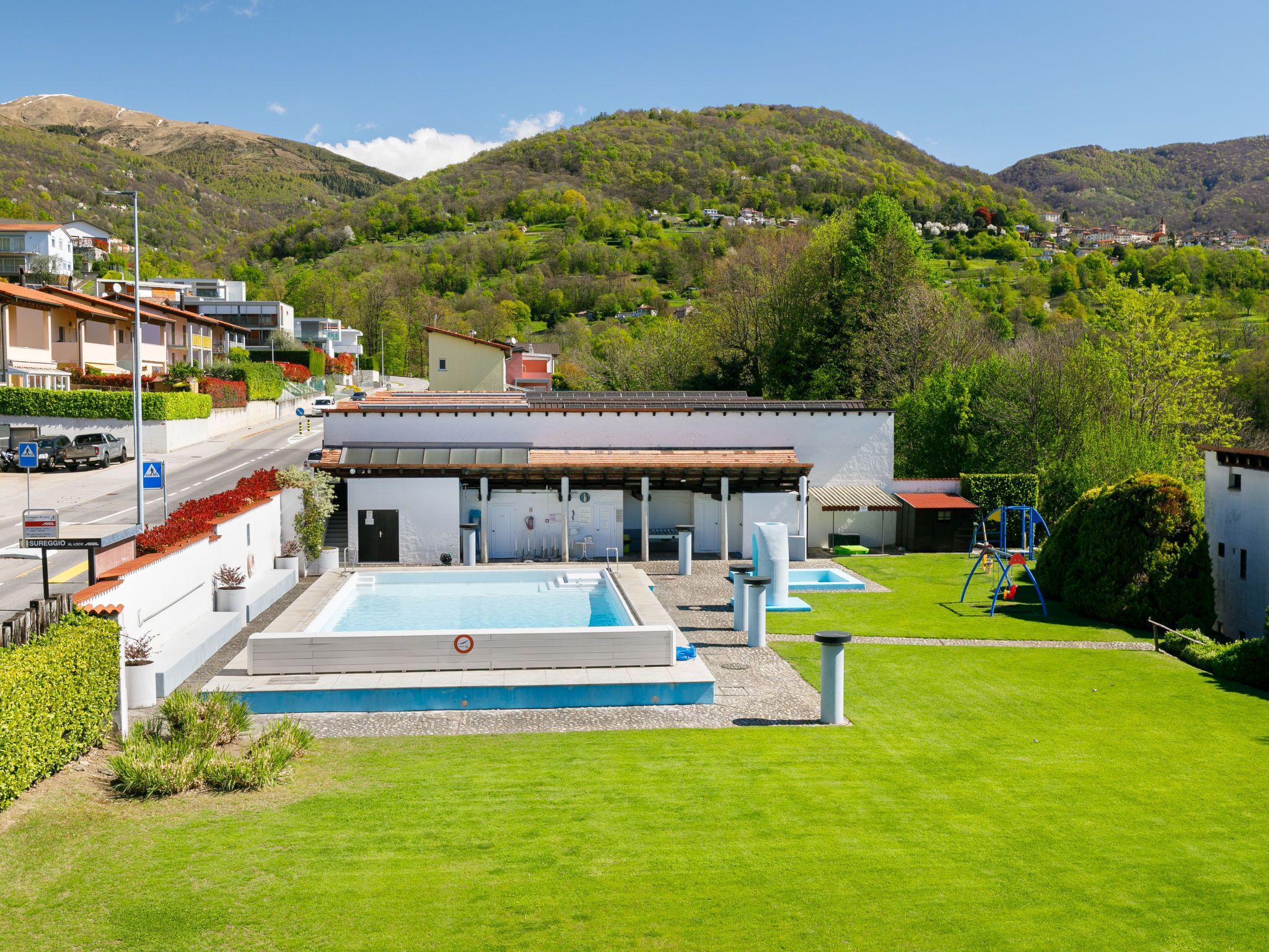 A modern building with a swimming pool, surrounded by a well-maintained lawn and a view of a hilly landscape.