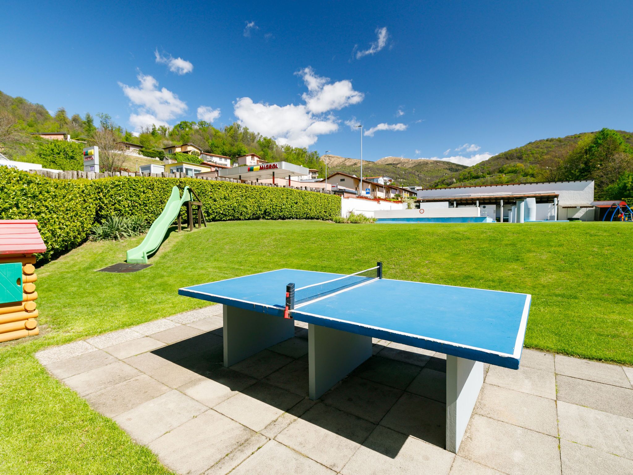 Une table de ping-pong se trouve dans un espace verdoyant, avec une aire de jeux et des bâtiments en arrière-plan.