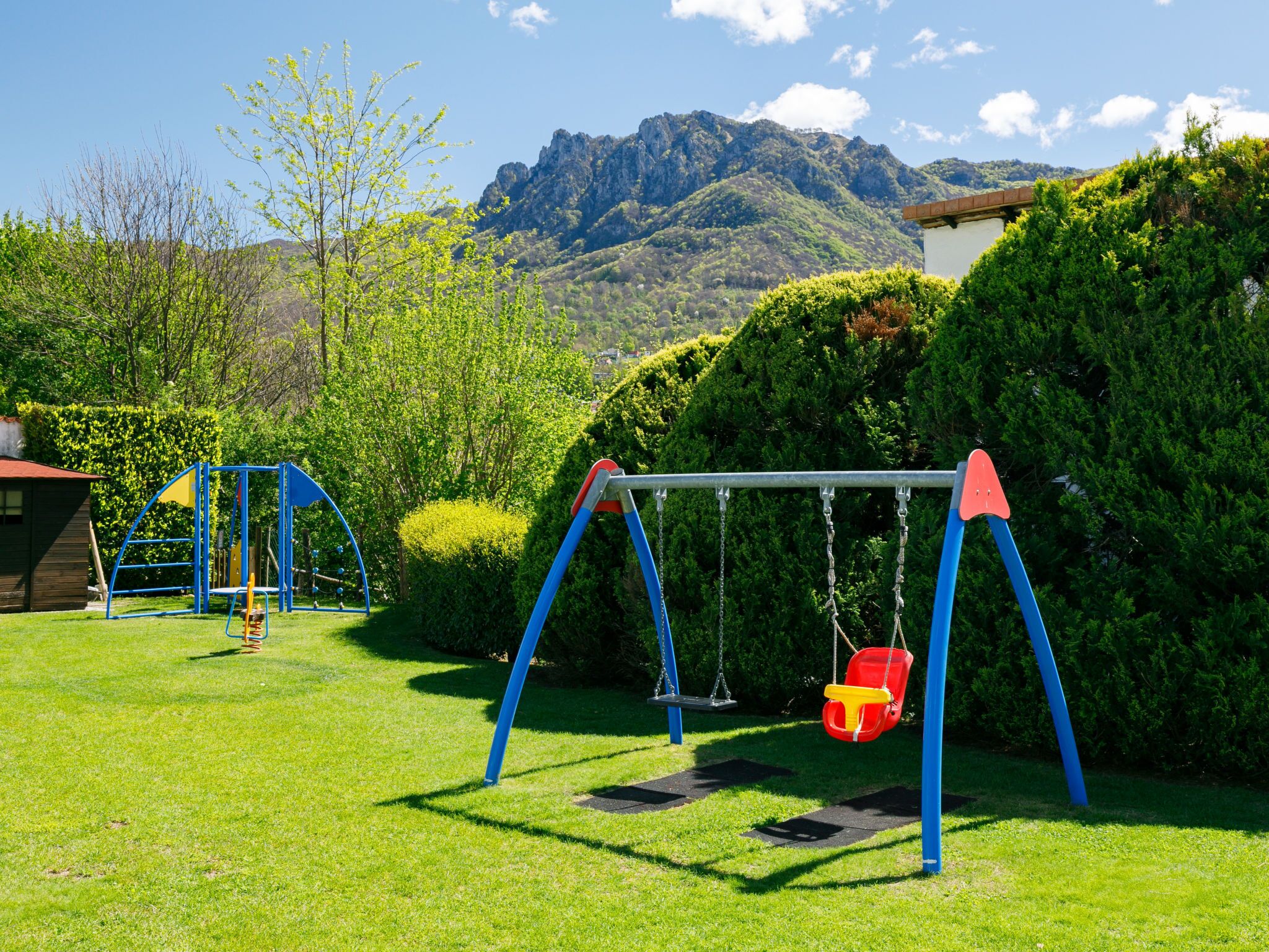 A playground with swings and a slide, surrounded by green grass and trees, with mountains in the background.