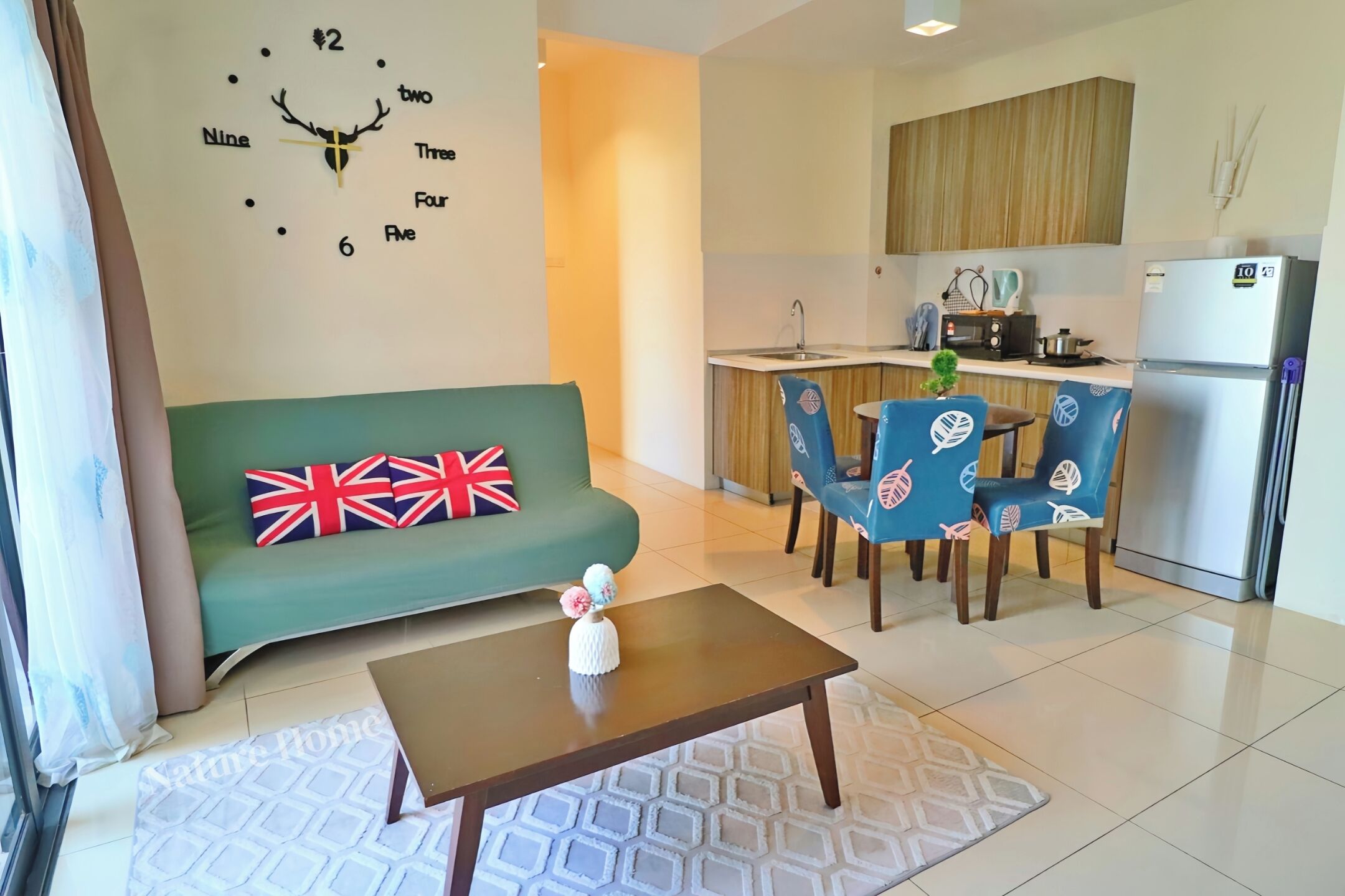 A living room with a green sofa, a coffee table, and a wall clock. There is a kitchen area with a refrigerator, microwave, and sink.