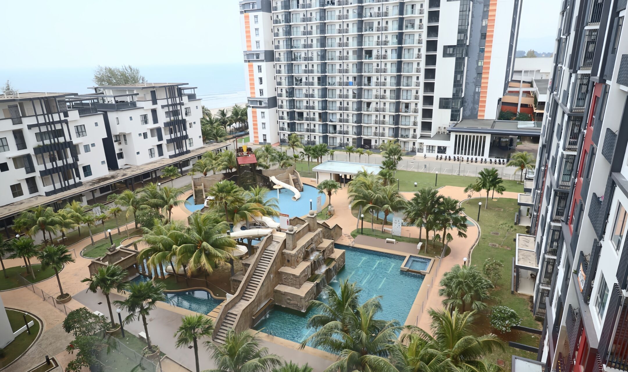 A high-rise building with multiple balconies, a swimming pool with a waterfall feature, and palm trees.