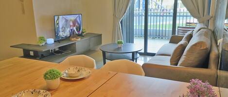 Una sala de estar con televisor, sofá, mesa de comedor y vista a un parque infantil a través del ventanal.