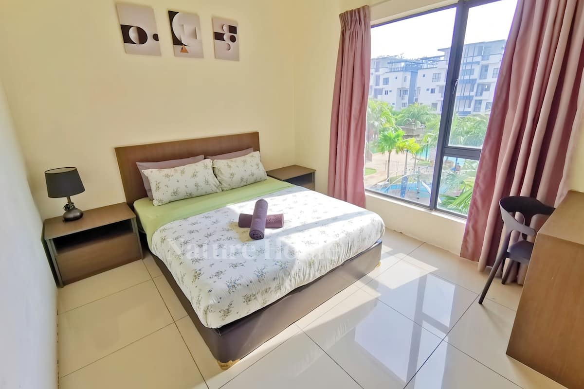 Une chambre d’hôtel avec un lit, une table de chevet, une lampe, une fenêtre avec des rideaux et une vue sur les bâtiments et la verdure environnante.